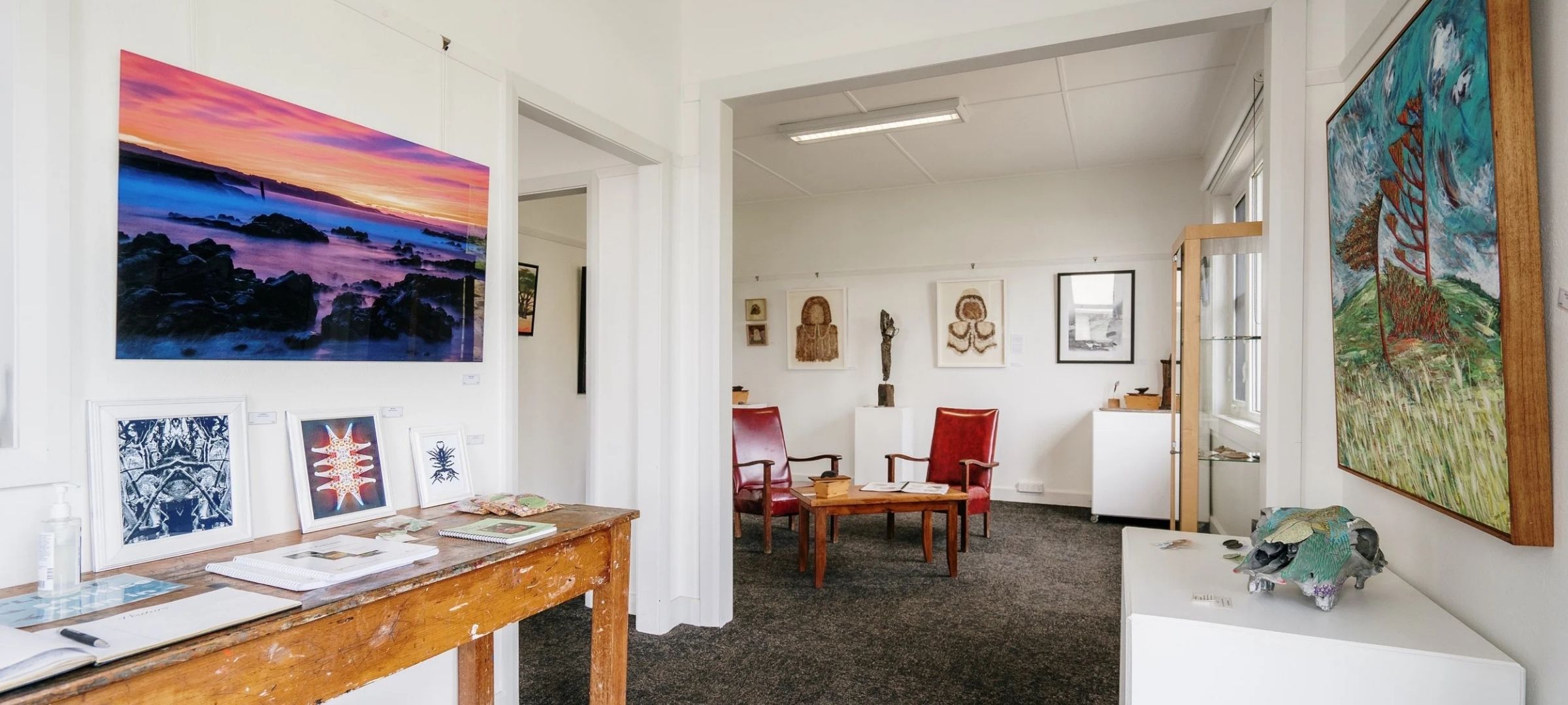 Interior of King Island Gallery, artwork and prints on white walls, red armchairs and a wooden table through to the next room.
